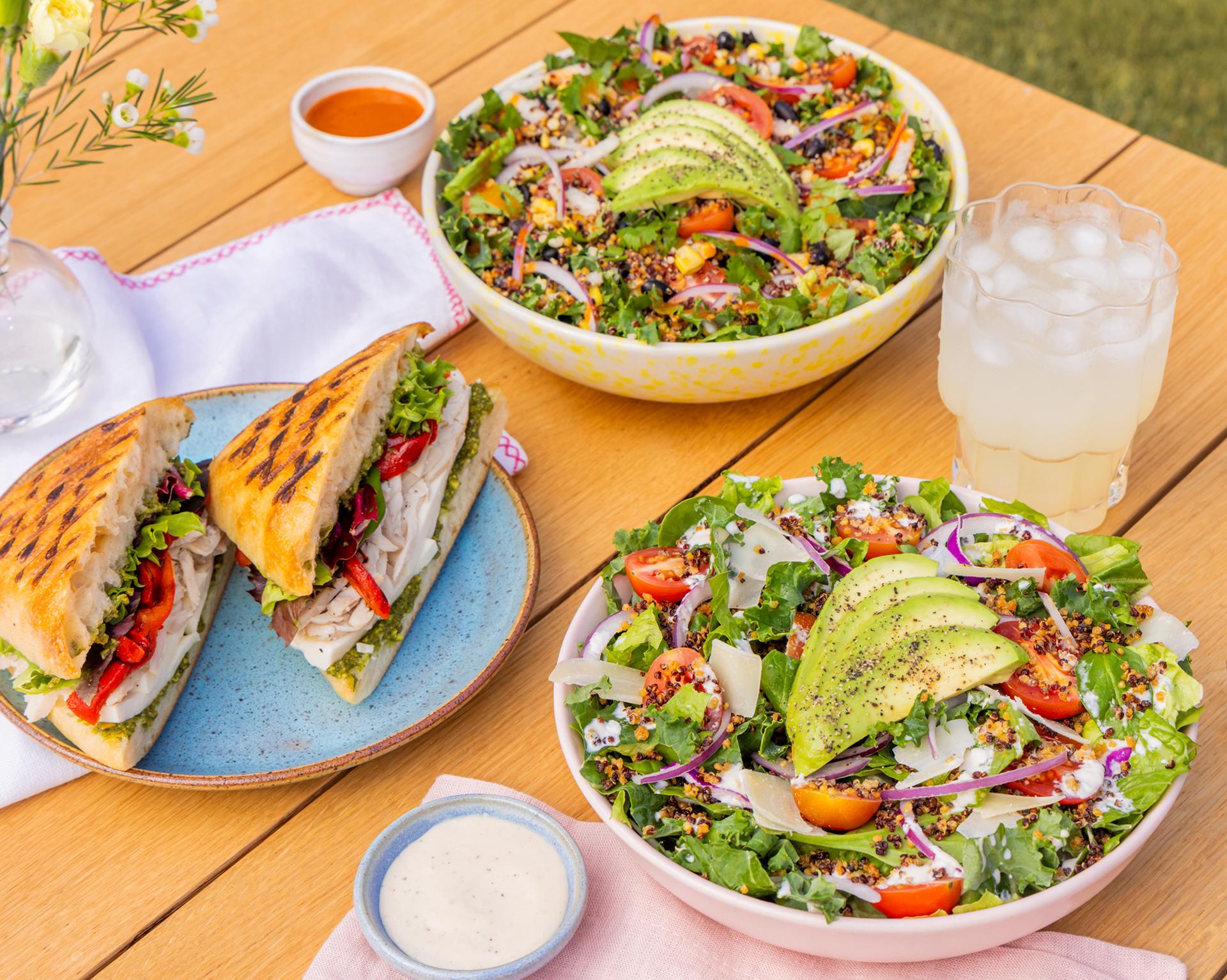 Fresh salads with sliced avocado, a grilled sandwich with turkey and veggies, a glass of iced drink, and dipping sauces on a wooden table.