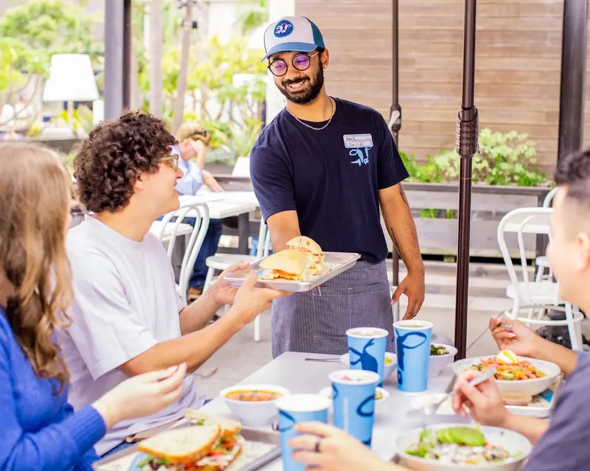 A server delivers a sandwich to a smiling group of four friends dining outdoors, with drinks and salads on the table.